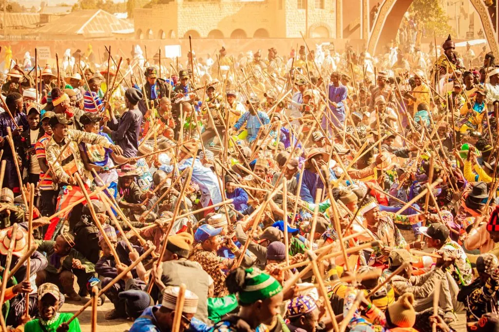 A vibrant outdoor scene of a traditional stick fighting ceremony with a large crowd participating energetically.
