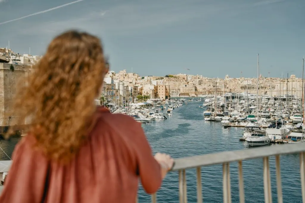Young woman enjoys a panoramic view of a marina in Senglea, Malta, with yachts and historic architecture.