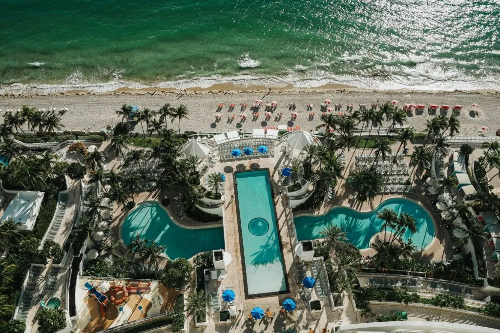 Aerial view of a beachfront resort with pools, palm trees, and sandy beach along the turquoise sea.