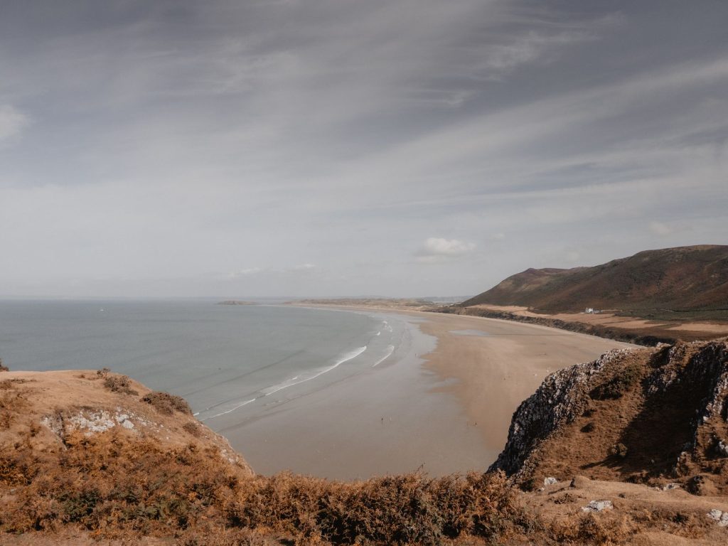 Expansive view of Rhossili Bay with sandy beach and lush cliffs under a cloudy sky.