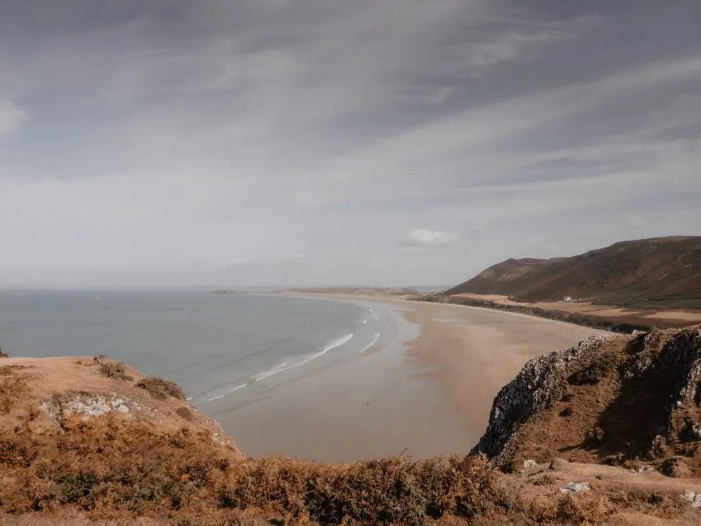 Expansive view of Rhossili Bay with sandy beach and lush cliffs under a cloudy sky.