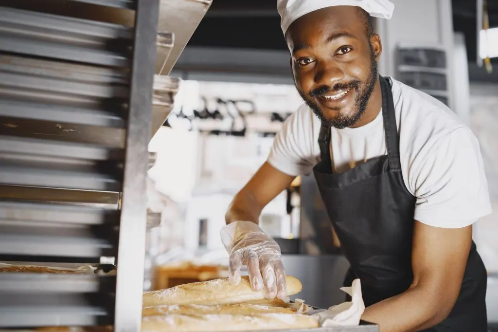 Cheerful male baker showcasing freshly baked baguettes in a bakery setting.