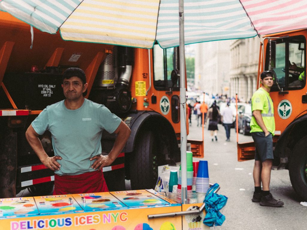 New York City street vendor selling ices beside construction trucks on a vibrant summer day.