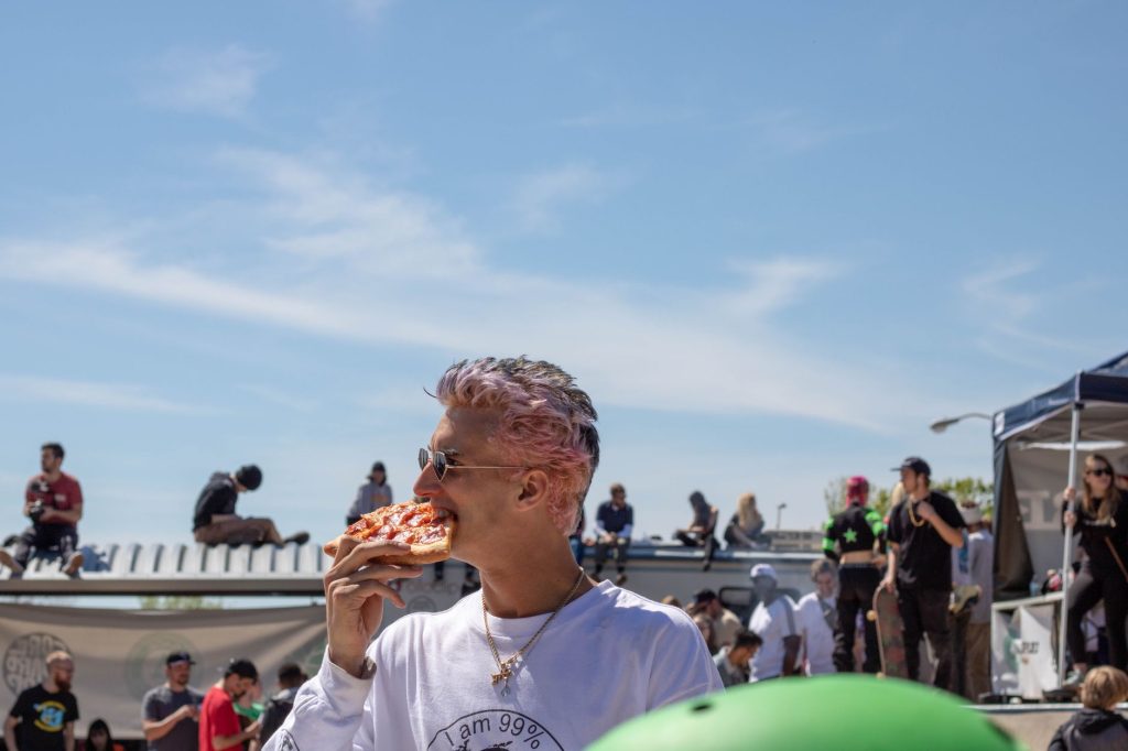 A man with dyed hair enjoys a pizza slice at an outdoor event under a clear summer sky.