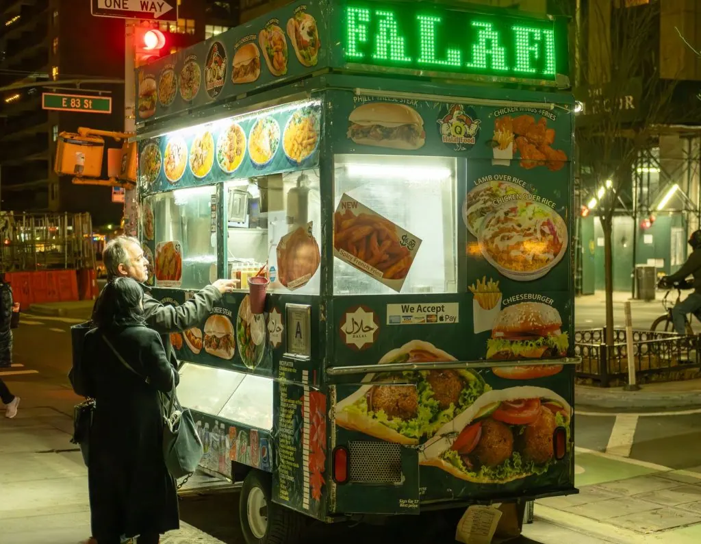 Vibrant street food scene in NYC with people ordering from a halal food cart at night.