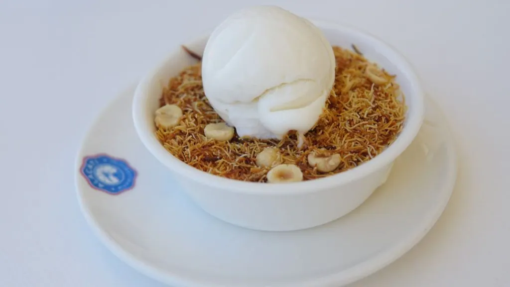 Close-up of a knafeh dessert served in a bowl with a scoop of ice cream on top, garnished with nuts.