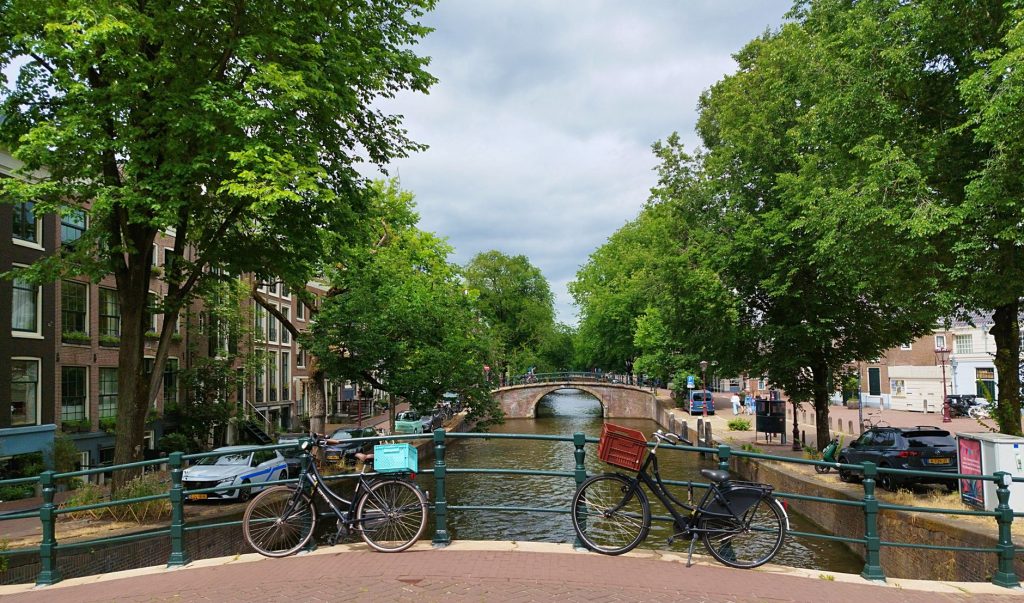 Scenic view of bicycles on a bridge overlooking a canal in Amsterdam, lined with trees and historic buildings.