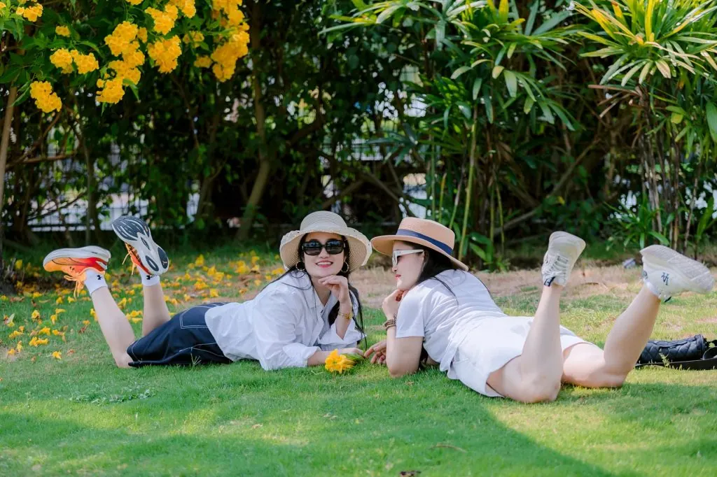 Two women in hats enjoy a sunny day on the grass, surrounded by vibrant yellow flowers.