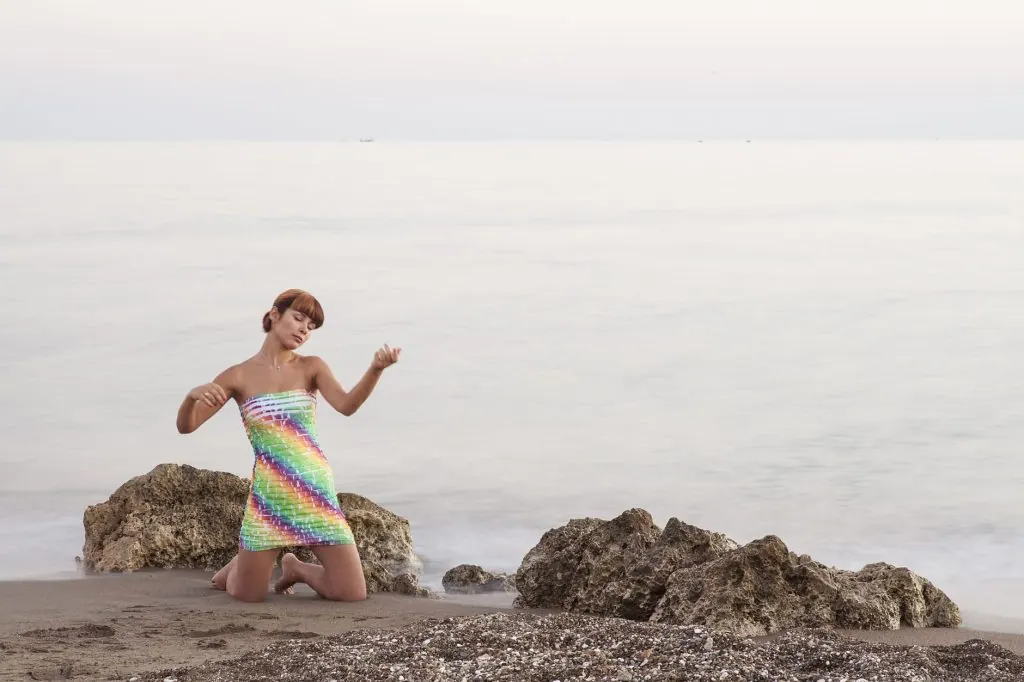 A young woman wearing a rainbow dress poses by rocks on a serene beach.