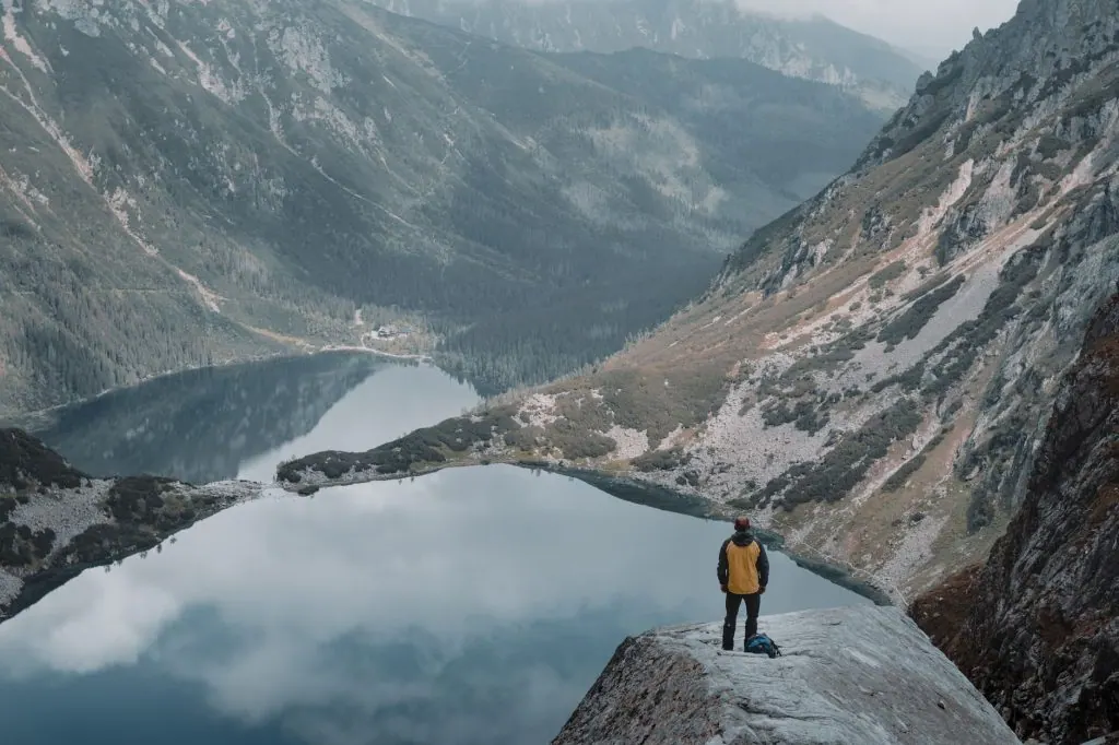 A hiker stands on a cliff overlooking a serene mountain lake surrounded by winter scenery.