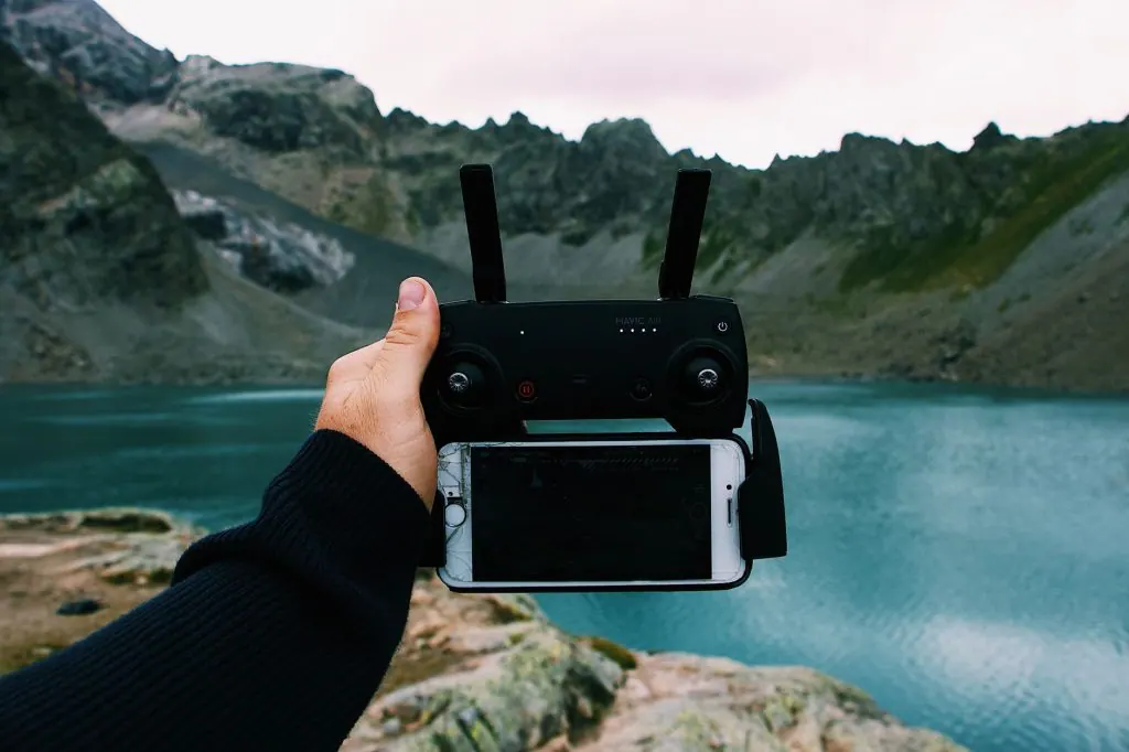 A hand holds a drone remote with a smartphone by a scenic mountain lake.