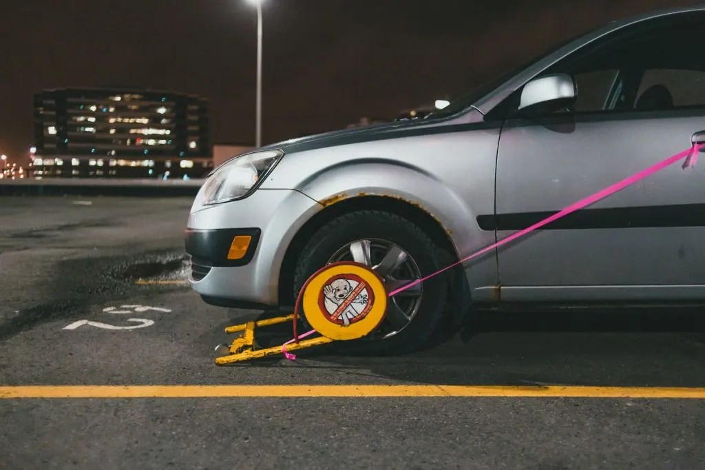 A silver car is immobilized with a wheel clamp in a dimly lit parking lot at night.