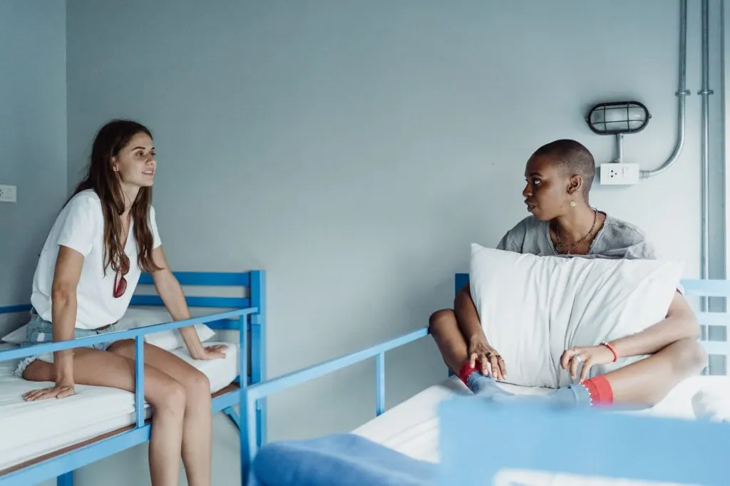 Two women sit and converse casually on bunk beds in a hostel room setting.