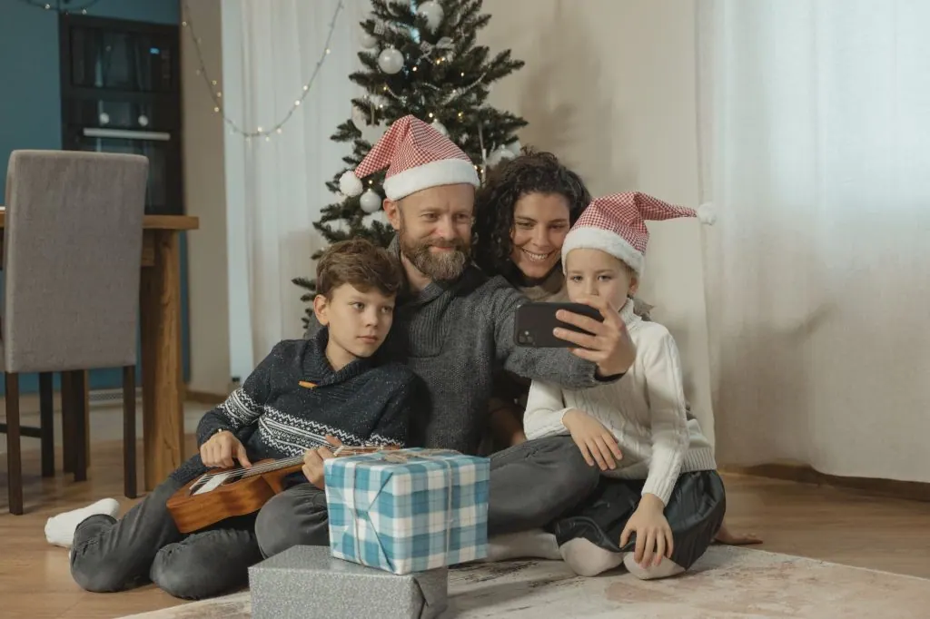 Happy family wearing Santa hats taking a selfie by the Christmas tree with gifts.