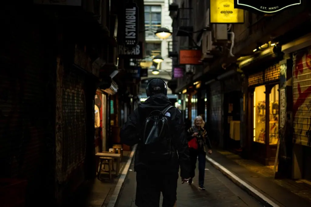 A person walking through a bustling Melbourne laneway in low light, surrounded by shops and graffiti.