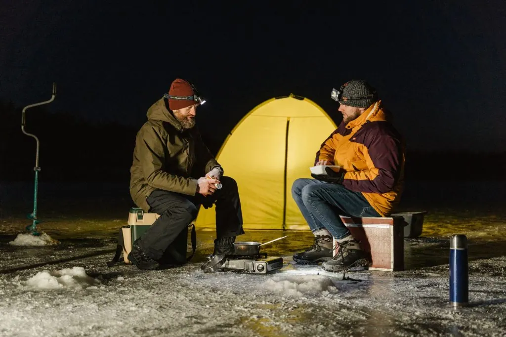Two men ice fishing at night with a tent on a frozen lake, enjoying a warm meal.