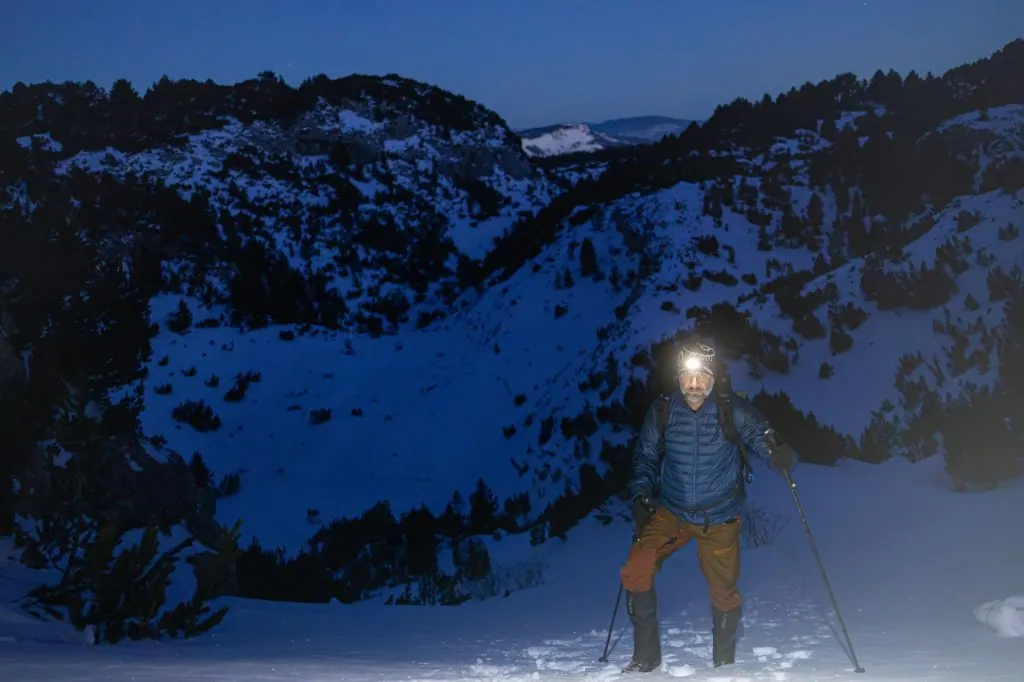 A solitary hiker climbs snowy mountains at night, guided by a headlamp.