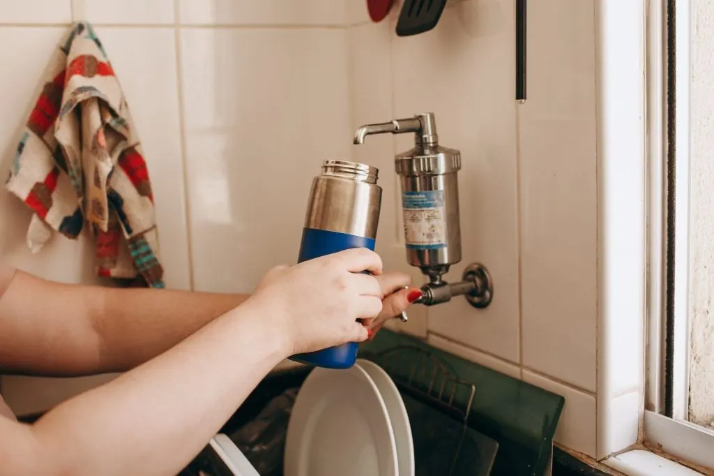 Close-up of hands filling a reusable water bottle at an indoor kitchen sink.