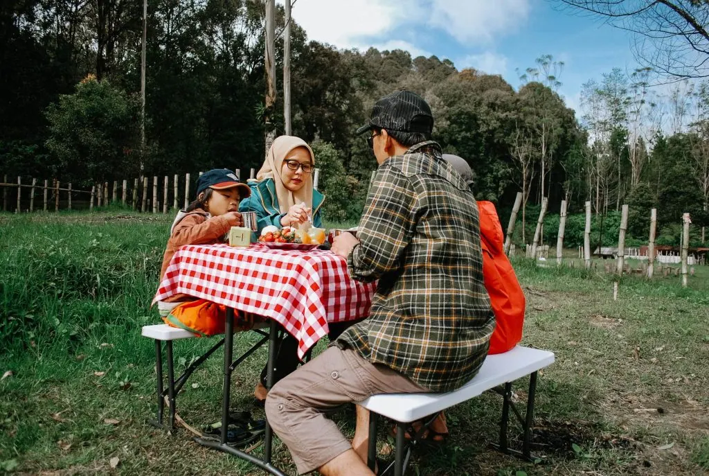 Family enjoying a meal at a picnic table outdoors in a scenic green park setting.