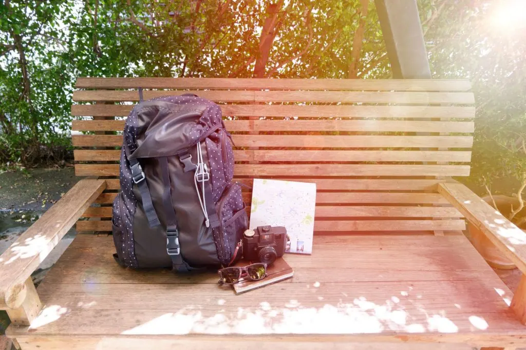 A backpack, camera, map, and sunglasses on a park bench with lens flare.