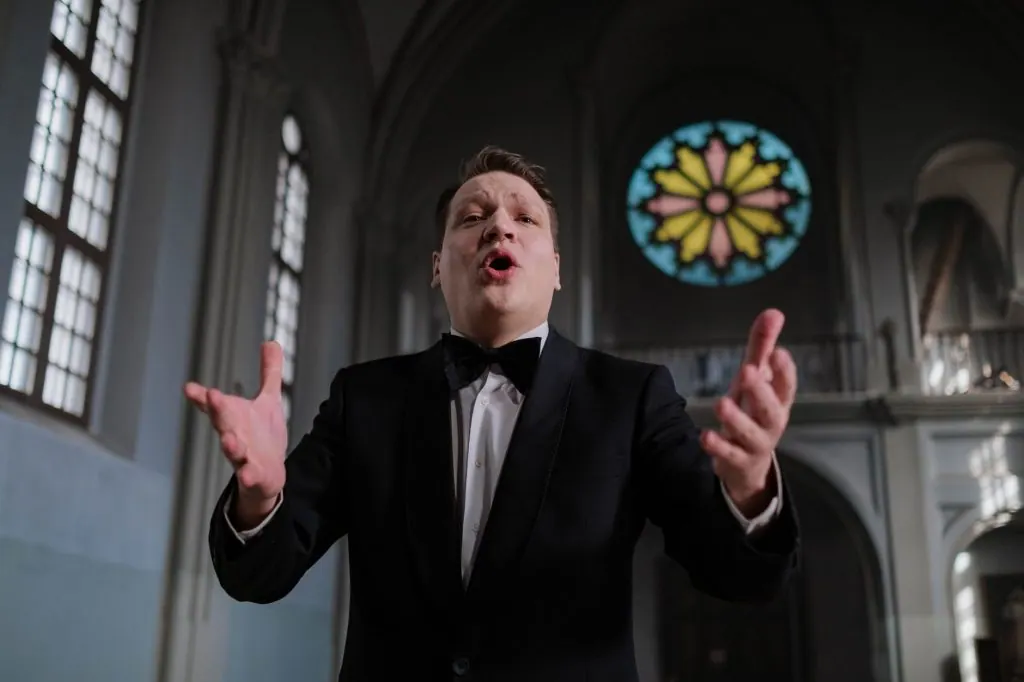 Opera singer performing passionately inside a church with stained glass window in the background.