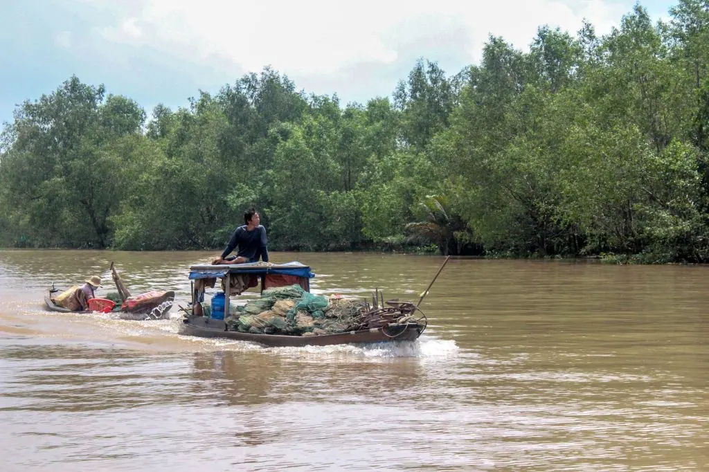 A man navigates a wooden boat on the Mekong River in Vietnam, surrounded by lush greenery.