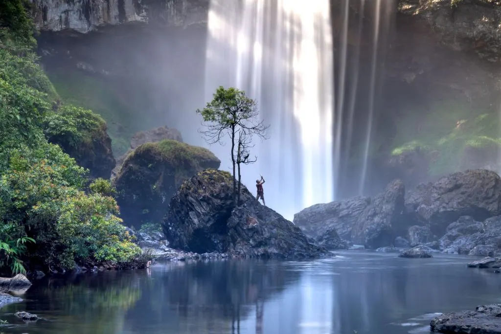 A person stands on a rock in front of a majestic waterfall, surrounded by misty, lush landscape.